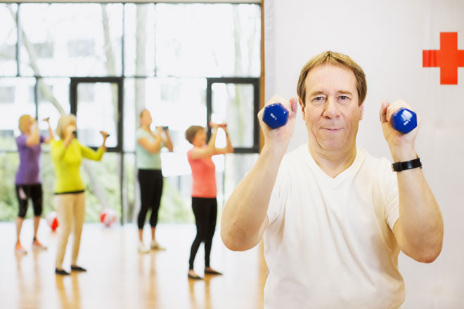 Ein Mann hebt mit jeder Hand eine Hantel. Im Hintergrund sieht man mehrere Personen, die Fitnessübungen machen