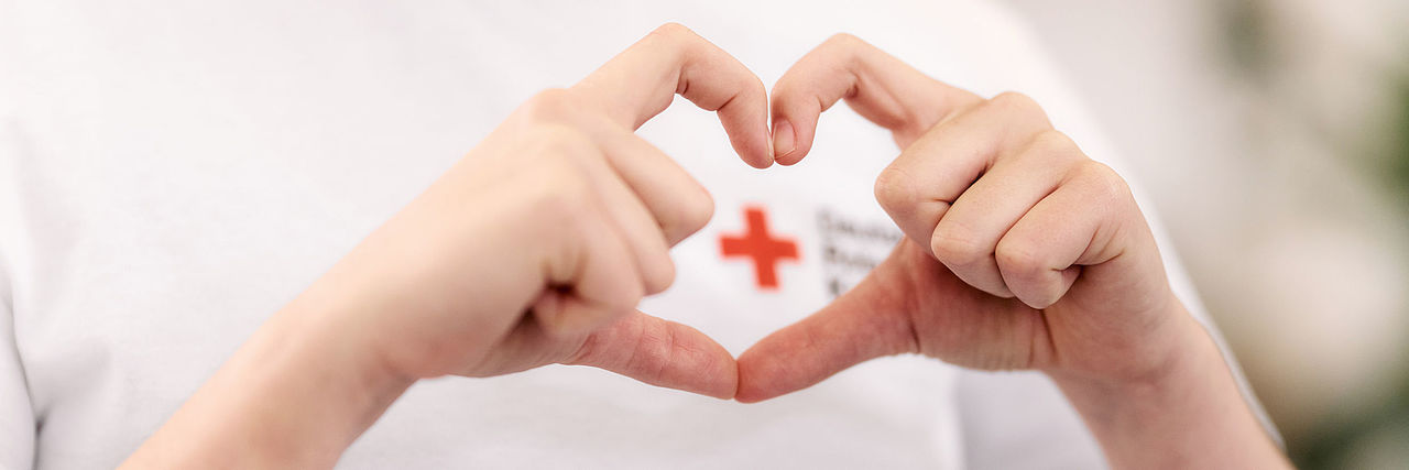 Finger formen ein Herz, DRK-Logo schaut aus dem Herz hervor Finger formen ein Herz, DRK-Logo schaut aus dem Herz hervor