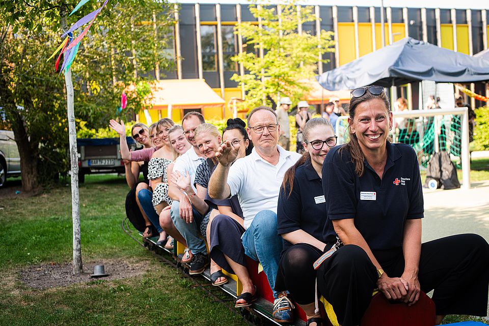 DRK Mitarbeitende sitzen auf einer Eisenbahn für Kinder zum Sommerfest. Alle Lächeln freundlich in die Kamera