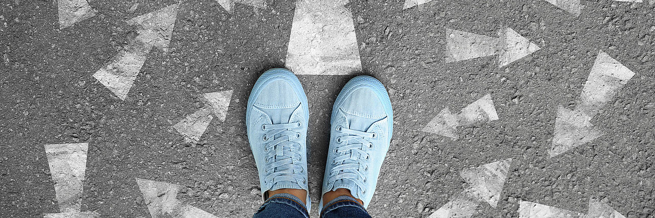 Füße mit Schuhen stehen auf Asphalt stehen. Pfeile weisen in unterschidliche Richtungen.