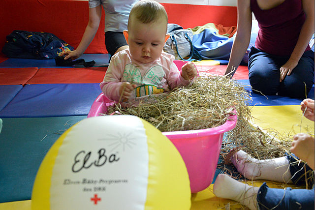 Kind sitzt in Schüssel mit Heu, davor liegt ein Wasserball mit Aufdruck ElBa