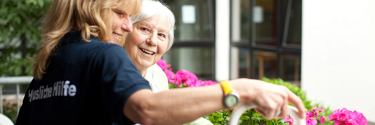 Pflegekraft gießt mit Seniorin Blumen auf dem Balkon