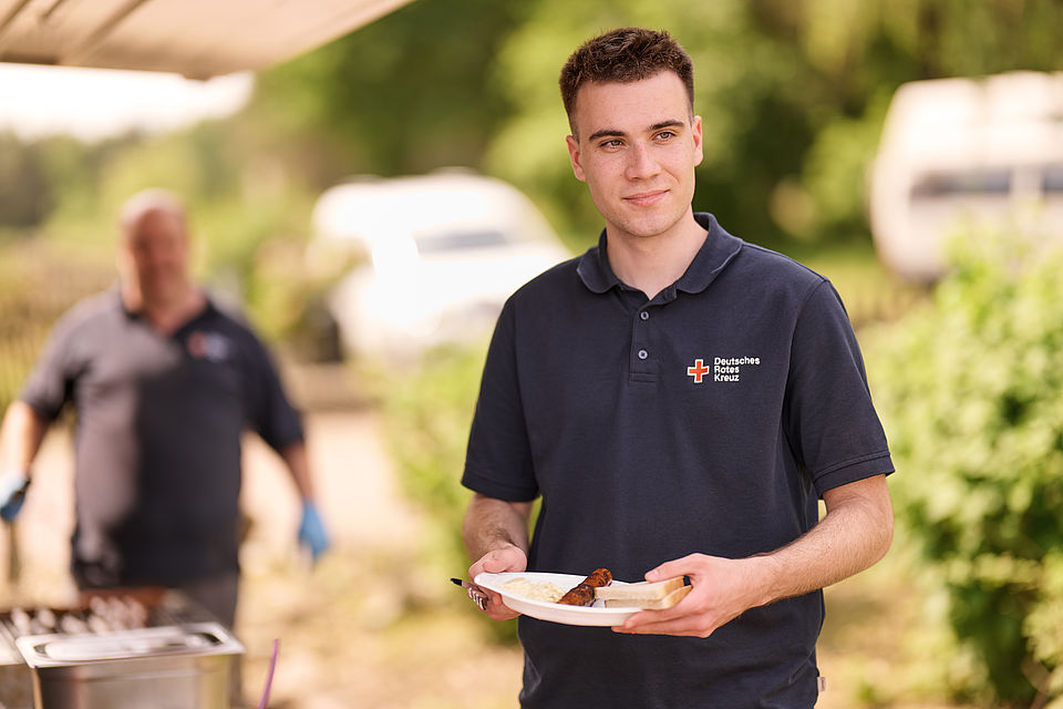 Ehrenamt beim DRK KV Diepholz: Training der Bereitschaft im Gelände. Verpflegungsgruppe beim Würstchen grillen. Ein Teilnehmer hat sich Verpflegung geholt. - Mai 2024