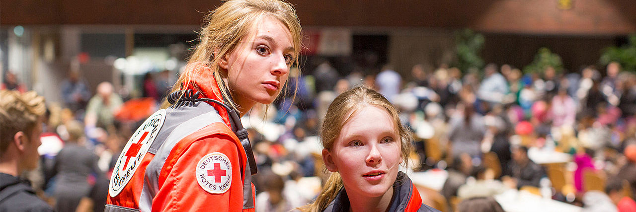 Kommen Sie in unser Ehrenamt! Zwei Frauen in Einsatzkleidung bei einer Veranstaltung mit vielen Menschen
