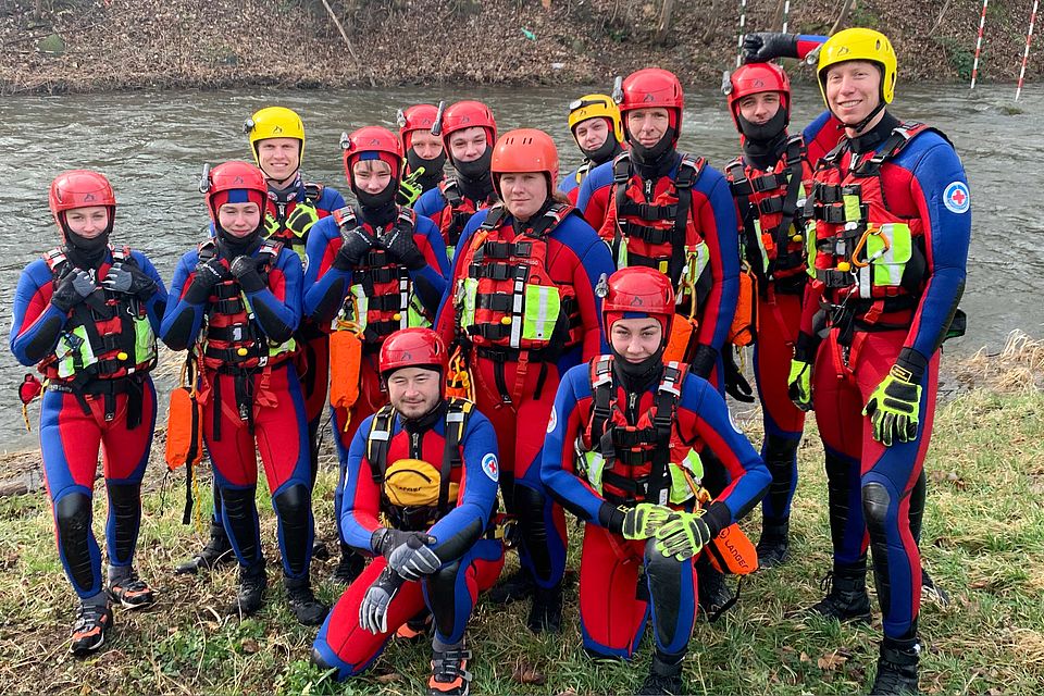 Junge Menschen mit Neoprenanzug und Helm bekleidet stehen an einem Fluss