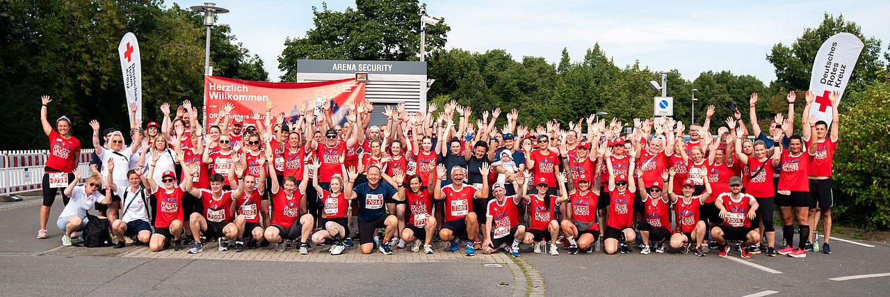 Viele DRK-Mitarbeitende vor dem Firmenlauf im Gruppenbild. Alle heben die Armen