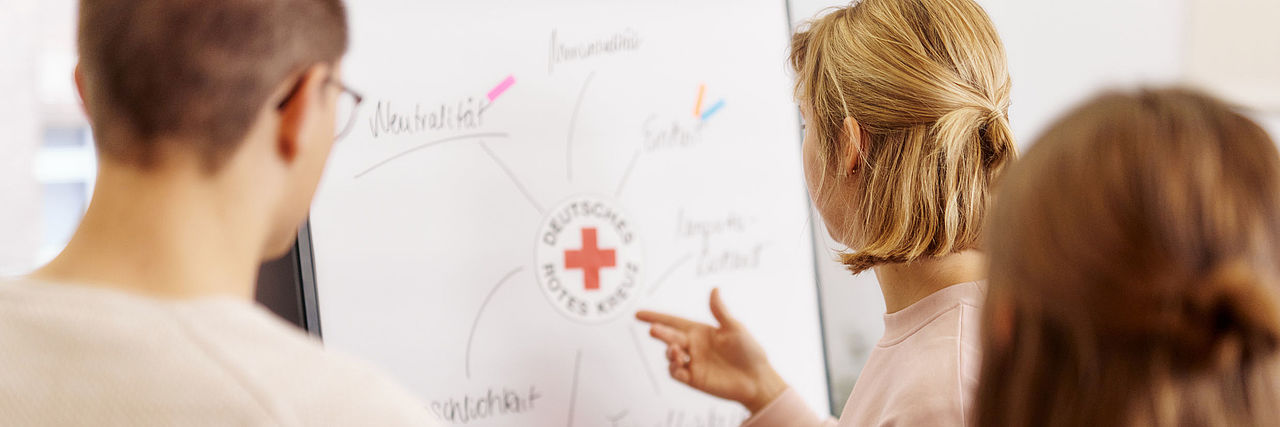3 junge Menschen stehen vor einem Flip Chart mit den Grundsätzen des DRK
