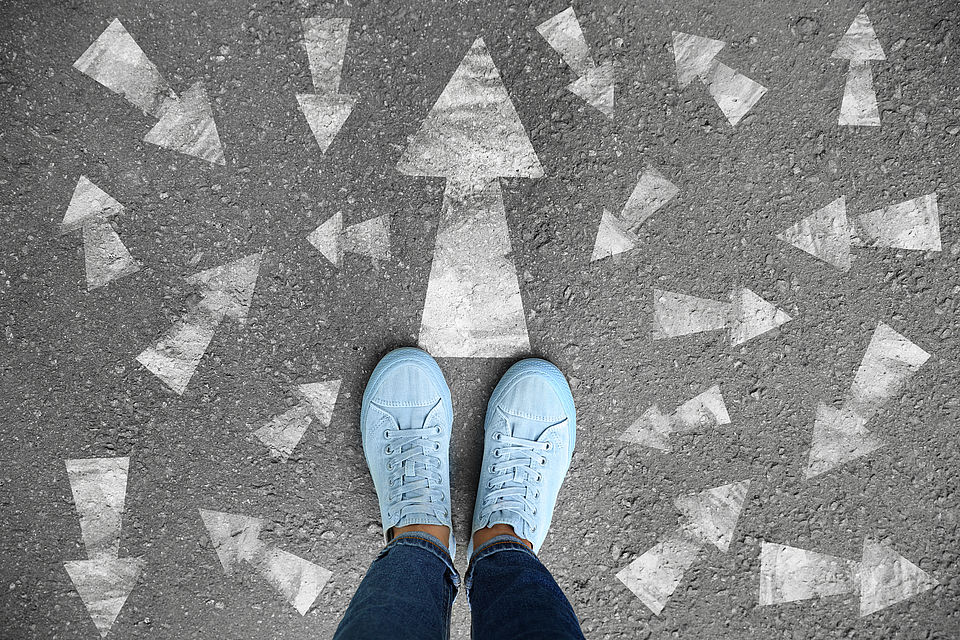 Blick auf Schuhe, die auf Asphalt stehen. Dieser ist mit Pfeilen in alle Richtungen bemalt