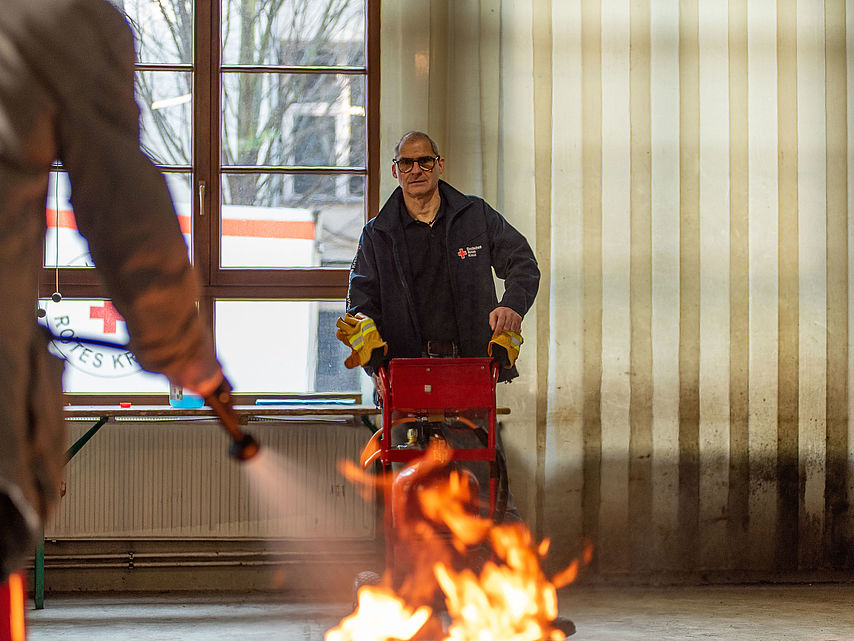 Ausbilder steht hinter Feuer am Brandsimulator, im vordergrund Person mit aktiviertem Feuerlöscher