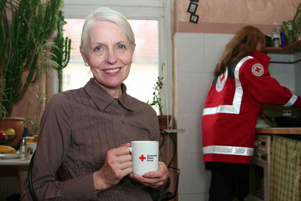 ältere Frau mit DRK-Tasse in der Hand, im Hintergrund eine Mitarbeitende des DRK