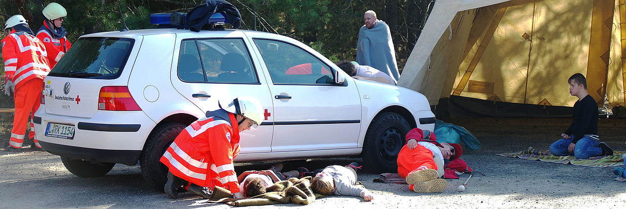 Verletzte liegen vor einem verunglückten Auto, Einsatzkräfte helfen