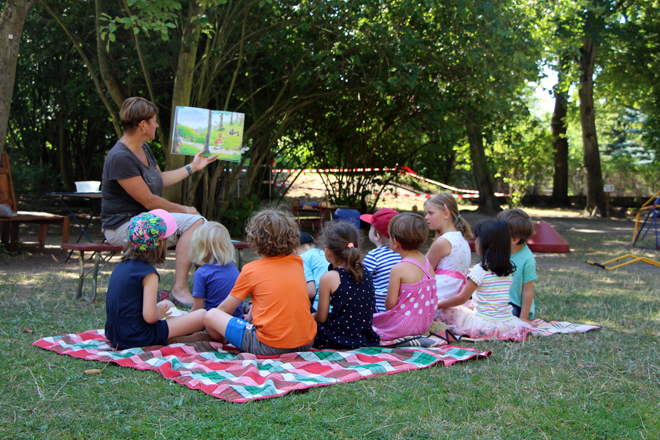 Mehrere Kinder sitzen auf einer Wiese, während ihnen eine Erzieherin ein Buch vorliest.